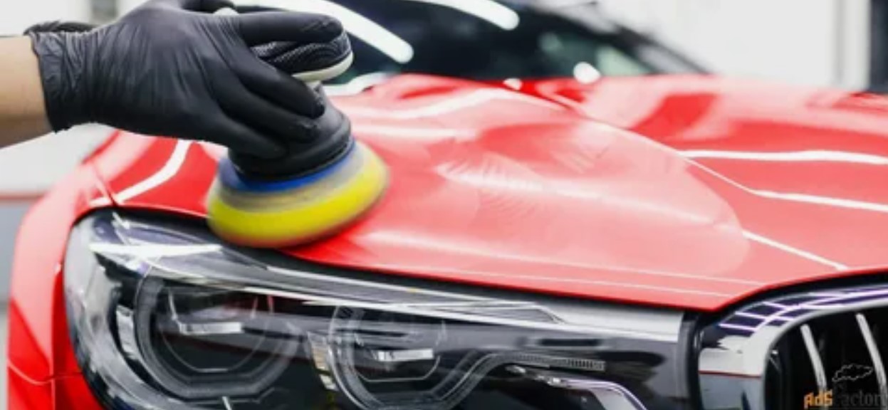 Uma mão com luva preta utiliza uma politriz para polir o capô de um carro vermelho, realçando o brilho da pintura.